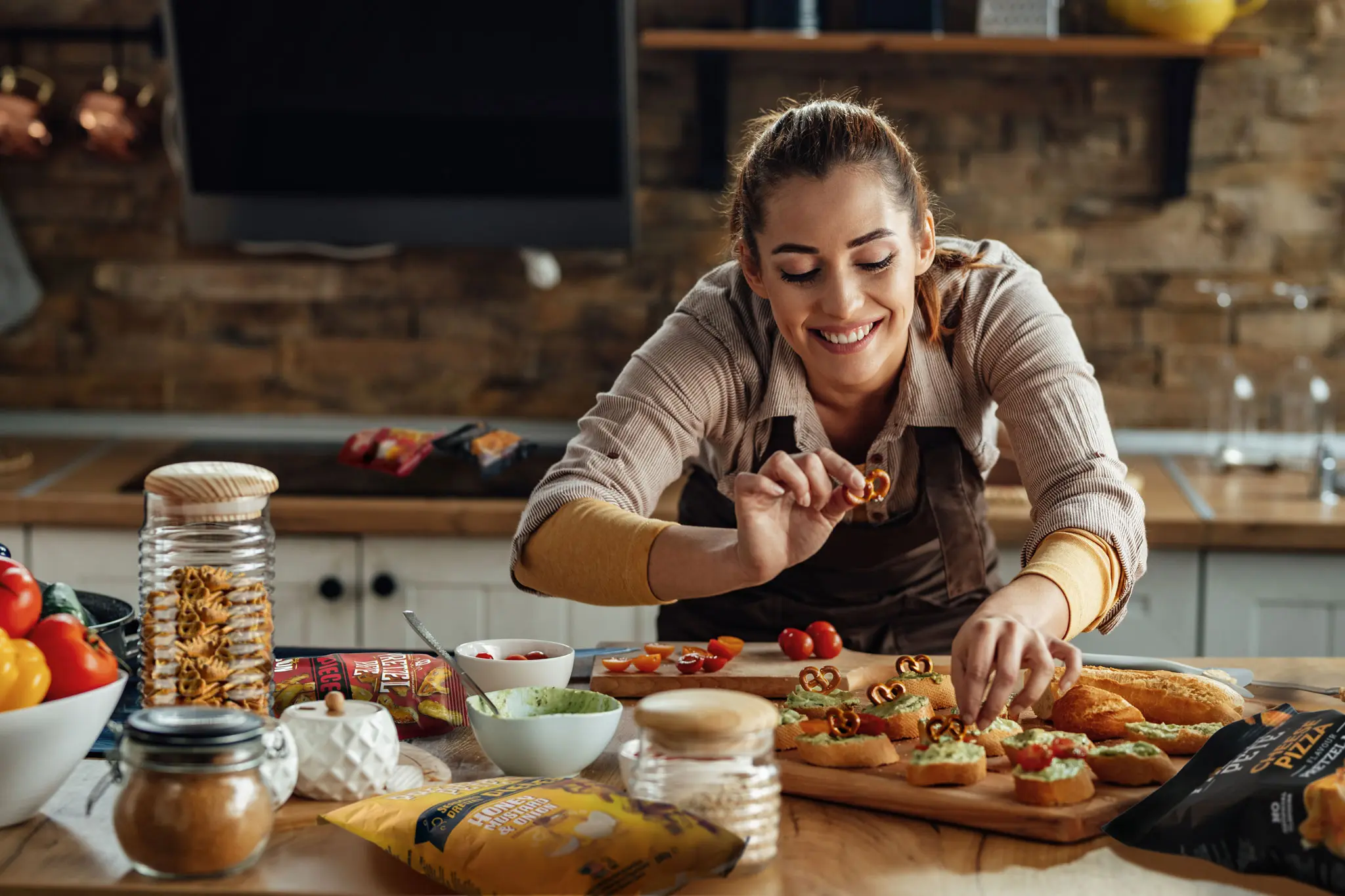 Gut gelaunte Frau kreiert in einer stilvollen, modernen Küche neue Rezeptideen mit Pretzel Pete Pieces. Auf dem Tisch- frisch belegte Bruschetta und kreative Snack-Kombinationen mit knusprigem Pretzel-Topping.
